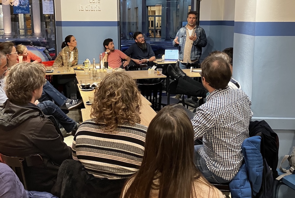 Foto del evento donde se ve a una persona presentando desde un rincól del café la diaria y el público escuchando alrededor de varias mesas unidas para la ocasión.