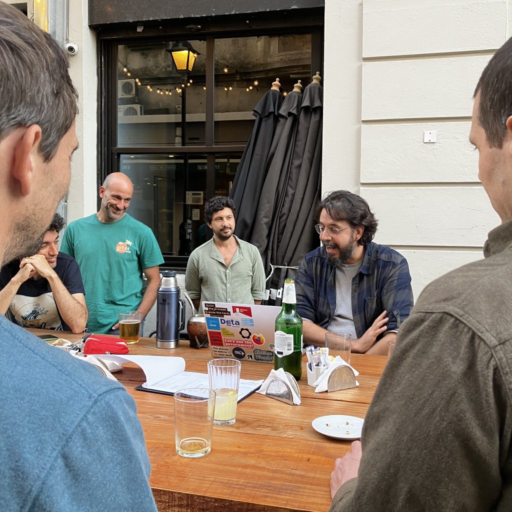 Sentados alrededor de una mesa alta, una personapresenta con su computadora, mientras el resto escucha y sonria.