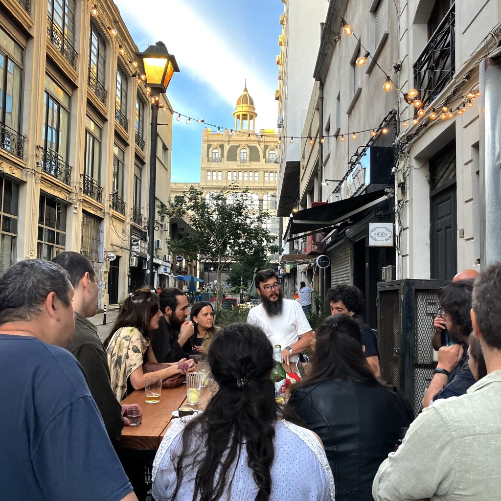 Vista del grupo alrededor de la mesa de café la diaria, con la peatonal Bacacay de fondo. Empieza a caer el sol y por encima de ellos está encendido unfarol estilo antiguo y una guirnalda de luces con bomitas pequeñas.
